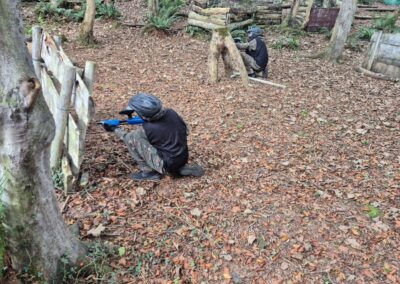 Two paintball players crouching behind barriers in a wooded paintball gallery.