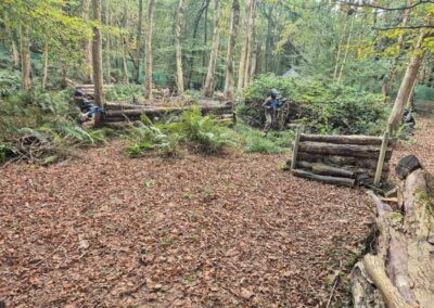 Paintball players in camouflage gear behind log barriers in a wooded area.