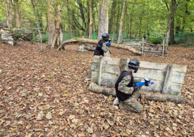 Two people playing paintball in a wooded area, aiming paintball guns behind wooden barriers.