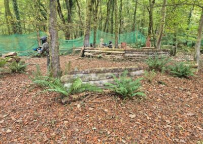Paintball players in a forest setting, taking cover behind wooden structures. Gallery.