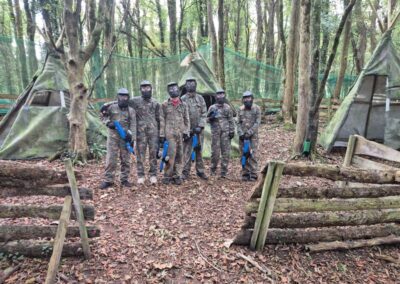 Group of paintball players in camouflage gear posing in a wooded paintball arena.