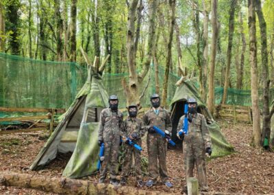 Paintball team in camouflage gear posing in a forest setting with teepee structures.