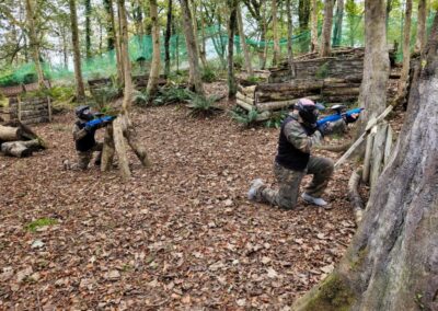 Two people playing paintball in a wooded area, aiming their markers during a game.