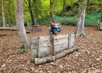 Person playing paintball in a forest, aiming their gun behind a wooden barrier.