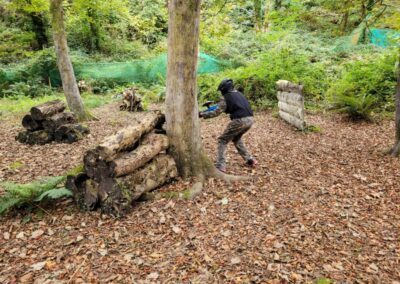 Paintball player aiming from behind a tree in a wooded paintball game.