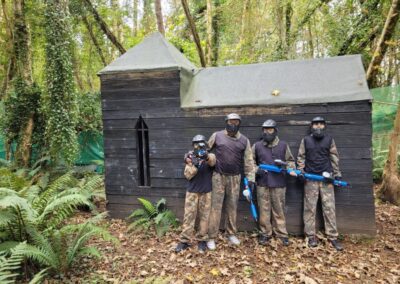 Group in paintball gear posing in front of a wooden structure, Gallery.