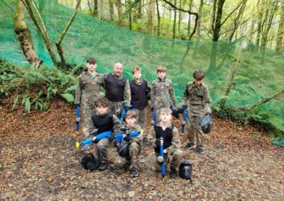 Group of kids and an adult in camouflage gear with paintball guns in a wooded area.