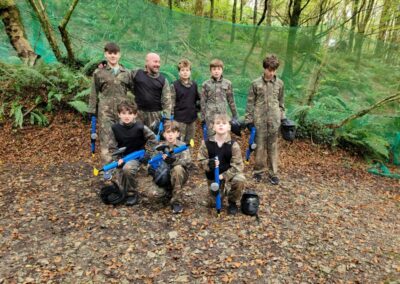 Group of boys and an adult in paintball gear pose for a photo in a wooded area.