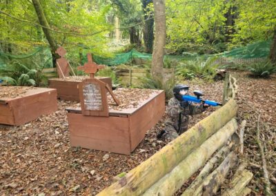 Paintball player aiming from behind wooden barrier in a wooded course with coffins. Text on coffin: 'Here lies John.'