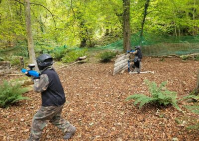 Players in paintball gear aiming paintball guns in a wooded area. Gallery.