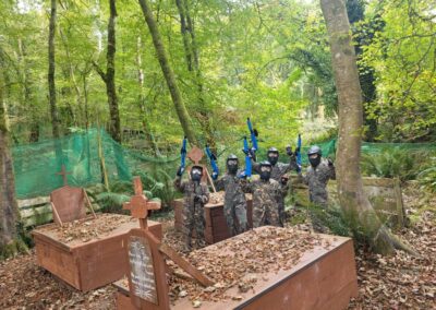 Paintball players in camouflage pose with guns near wooden coffins at a paintball field. Gallery.