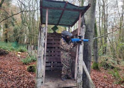 Paintball player in camouflage aims blue marker from wooden tower in the woods.